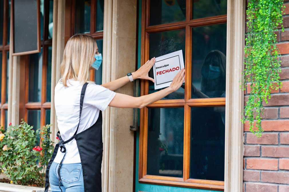 Funcionária de máscara e avental colando placa de "fechado" em porta de estabelecimento